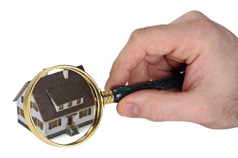 A hand holding a magnifying glass over a small model house.