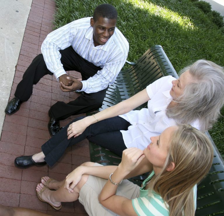 Three people conversed while sitting on a bench in a green outdoor setting.