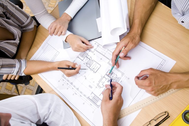 Hands discussing plans and diagrams on a table with pens and blueprints.