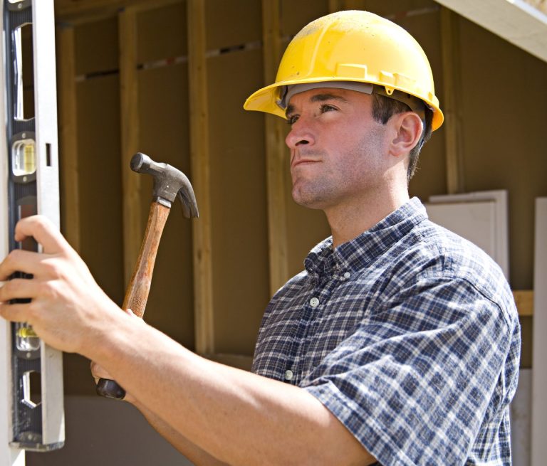 A construction worker uses a level, holds a hammer, and wears a hard hat.