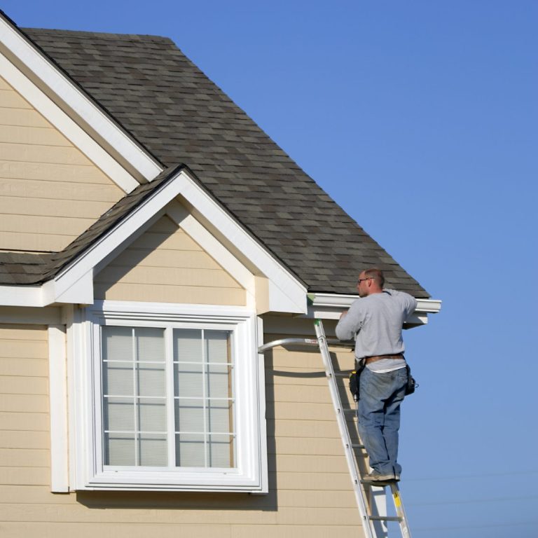 A man on a ladder repairing the eaves of a beige house under a clear blue sky.