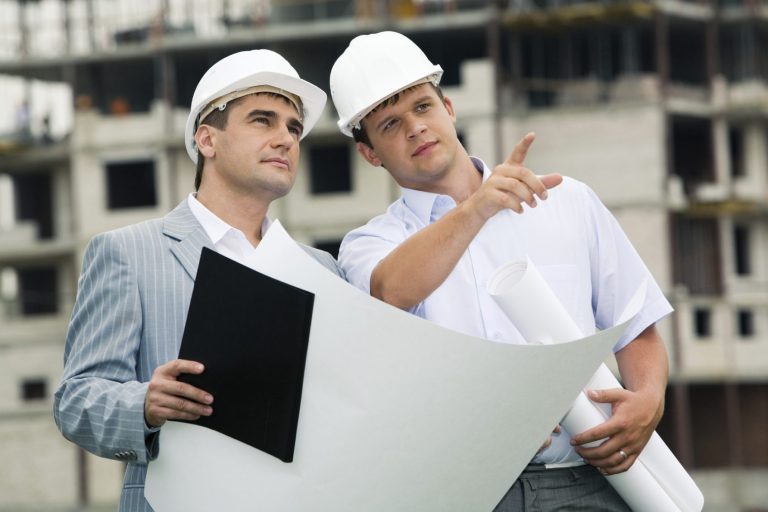 Two men in hard hats examine blueprints at a construction site.