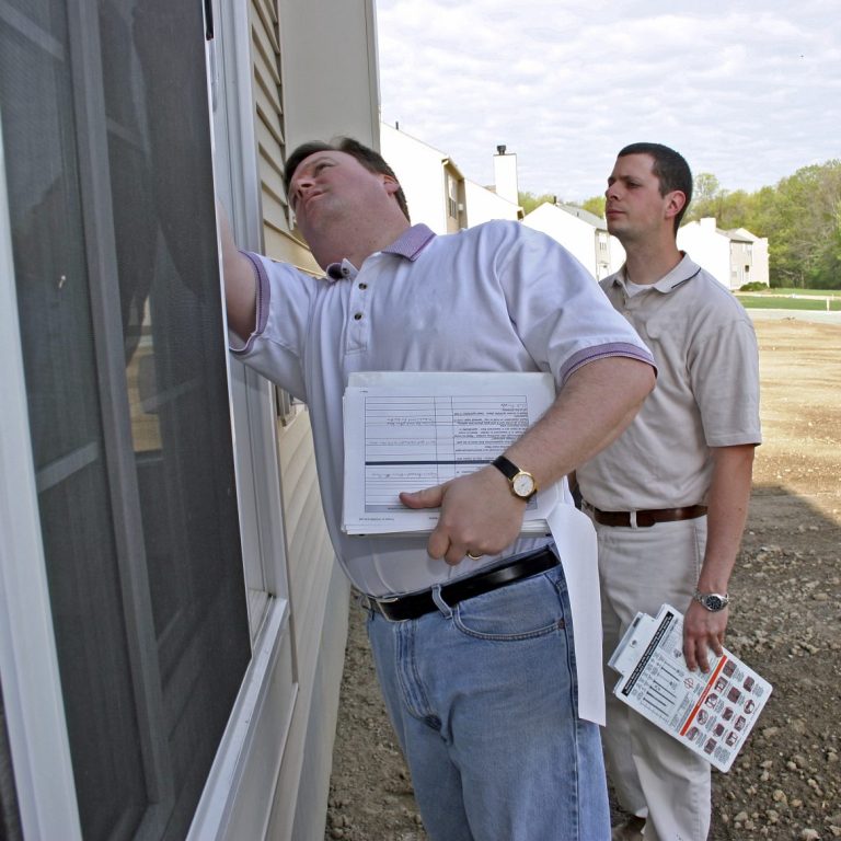 Two men inspecting a building, one examining a window and the other holding documents.