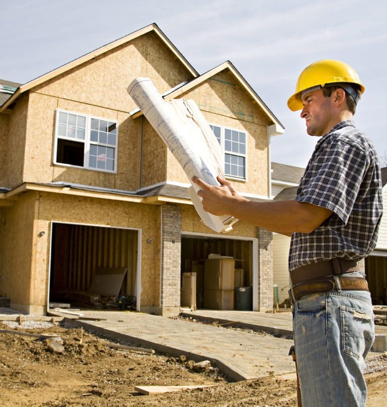 A construction worker in a hard hat reviewing blueprints in front of a house under construction.