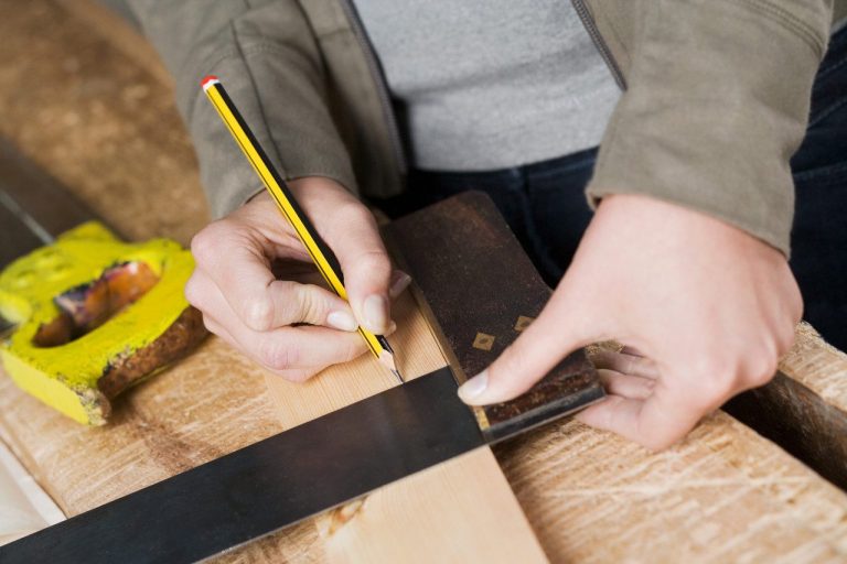Person measuring and marking wood with a pencil and square tool on a workbench.