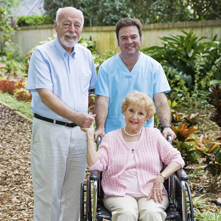 Two men in a garden surround a smiling elderly woman in a wheelchair.